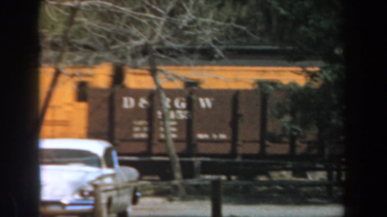 A white classic car sits in front of brown D&RGW train cars and trees.