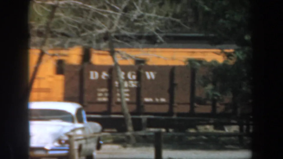 A white early-1960s car appears in front of D&RGW-marked train cars and trees at Knott's Berry Farm, California.