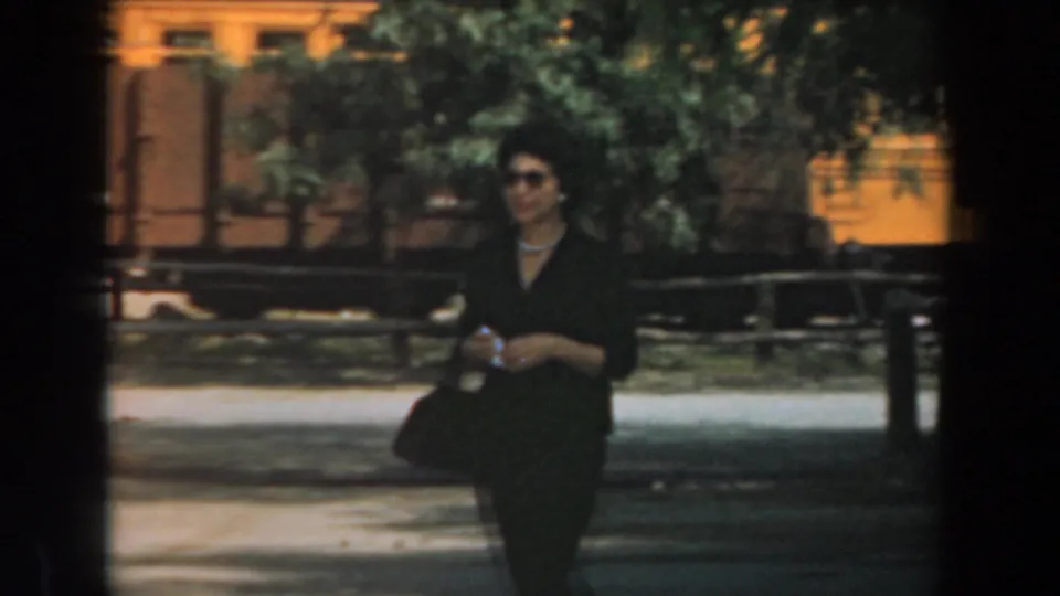 A well-dressed woman in sunglasses walks past trees and rail cars in this 1961 Knott's Berry Farm home-movie frame.