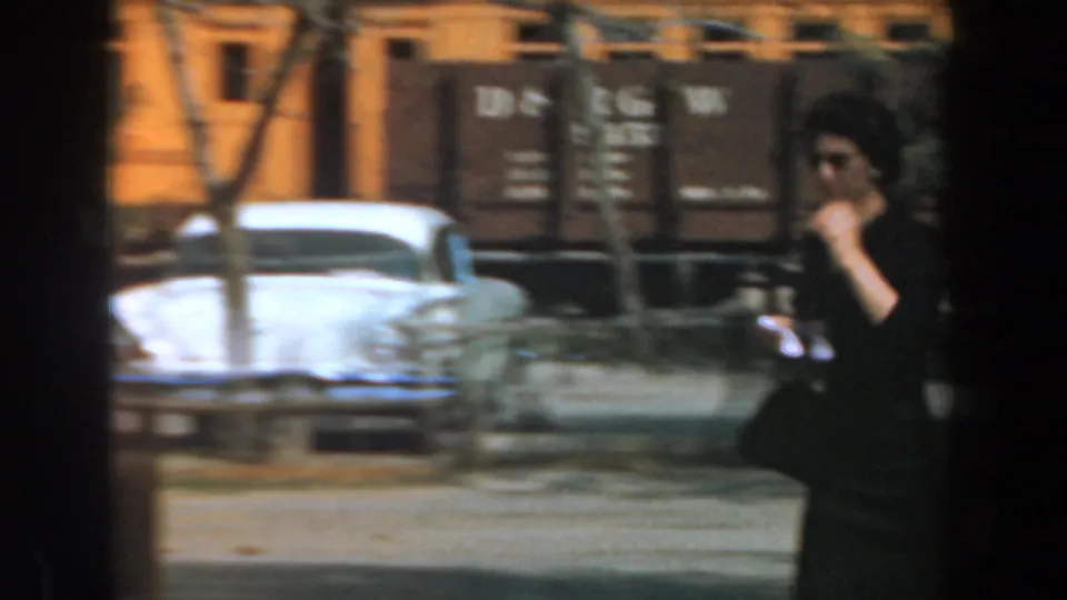 The woman continues walking past a white classic car while D&RGW rail cars move behind trees at Knott's Berry Farm in 1961.