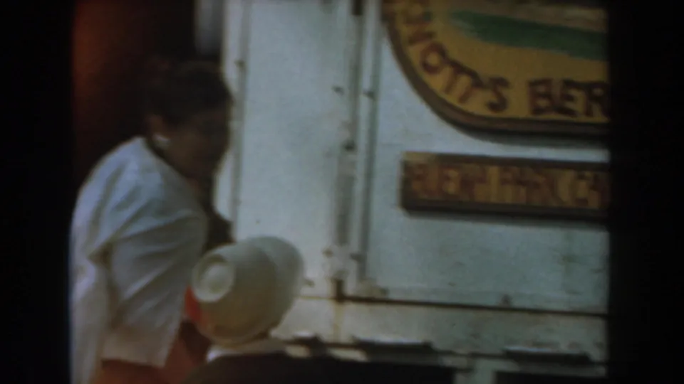 Home-movie frame showing a woman near the Knott's Berry Farm sign, with another visitor's white hat visible in the foreground.
