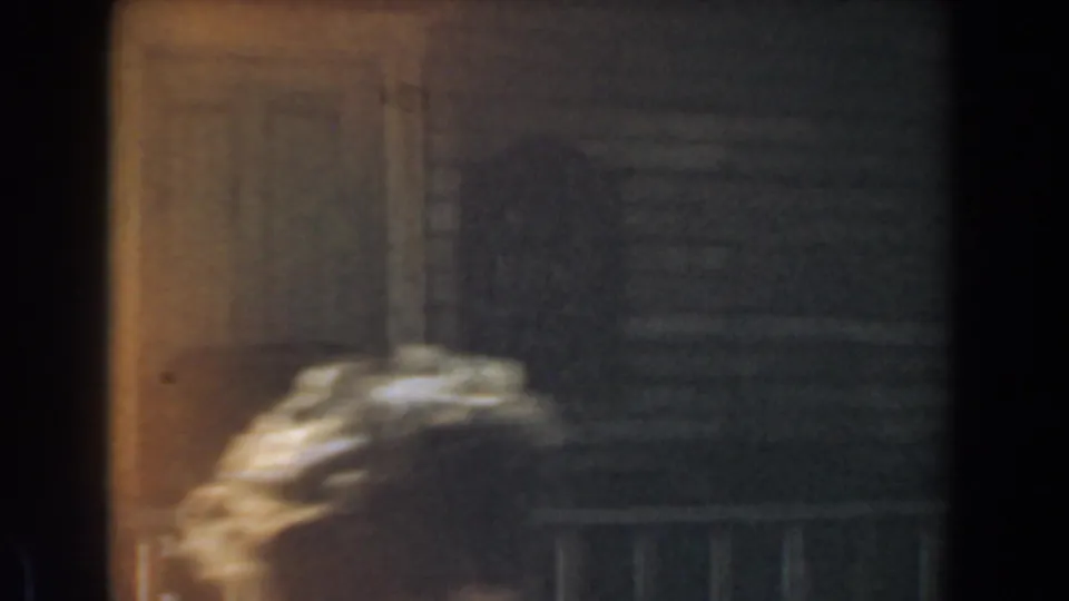 Grainy 1961 Knott's Berry Farm frame showing a dark rustic exterior, porch rail, and a bright blurred shape at lower left.