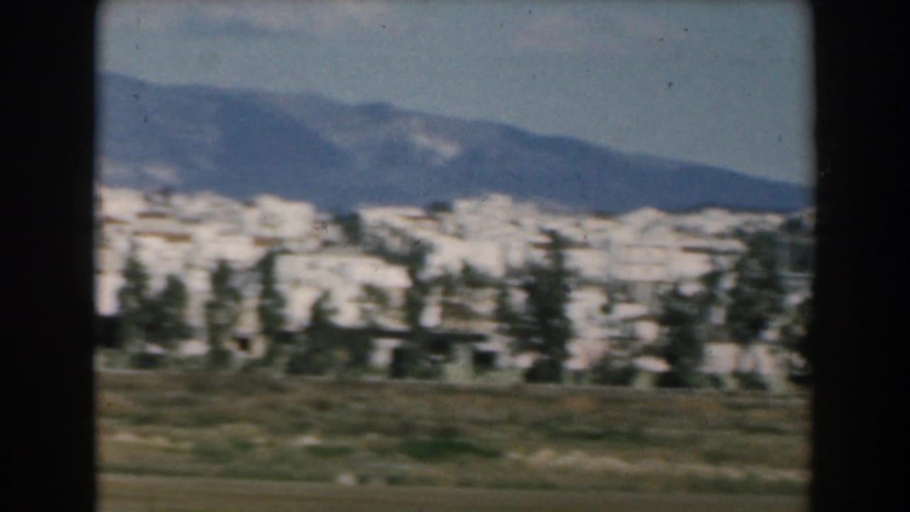 1962 Greece home-movie frame showing white town buildings, scattered trees, open ground, and mountains in the distance.