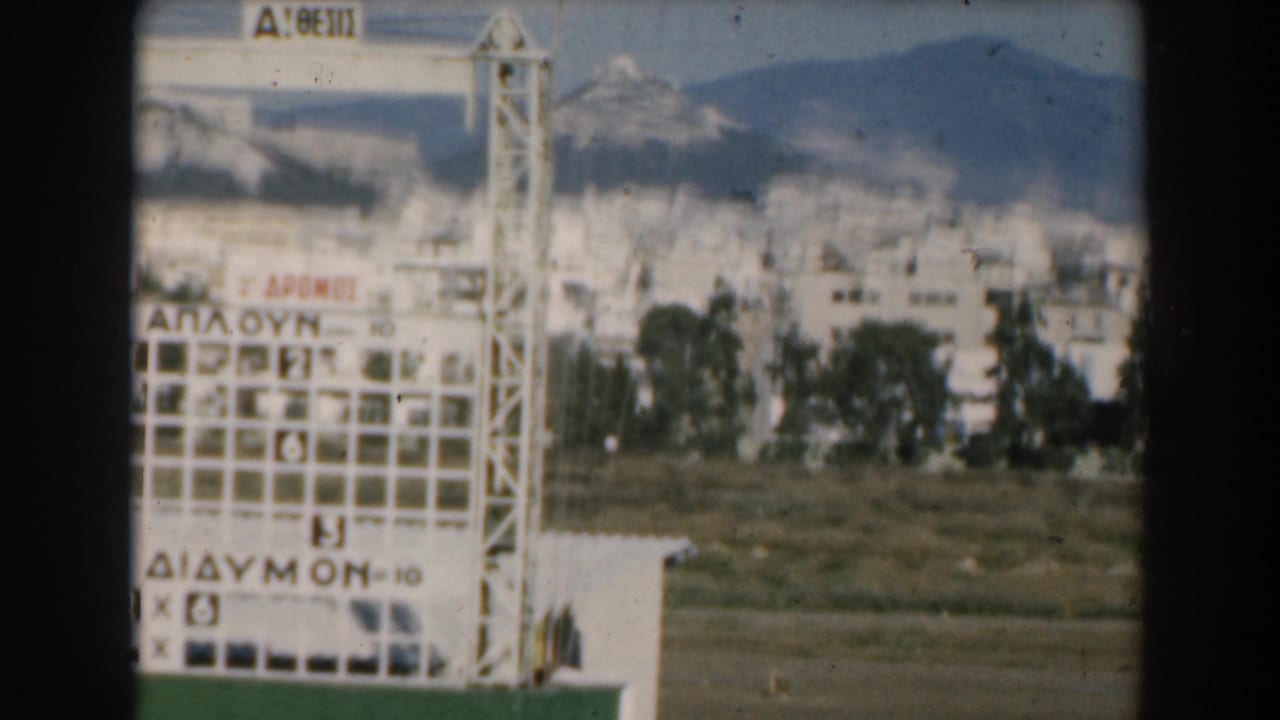 1962 Greece frame with a scoreboard or sign structure carrying Greek text, with town buildings and mountains behind it.