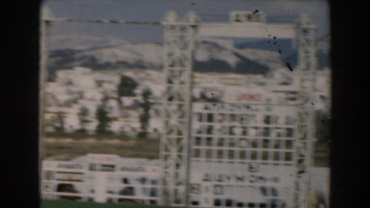 1962 Greece home-movie frame showing a passing scoreboard-like frame, a bus or coach, town buildings, and mountain slopes.
