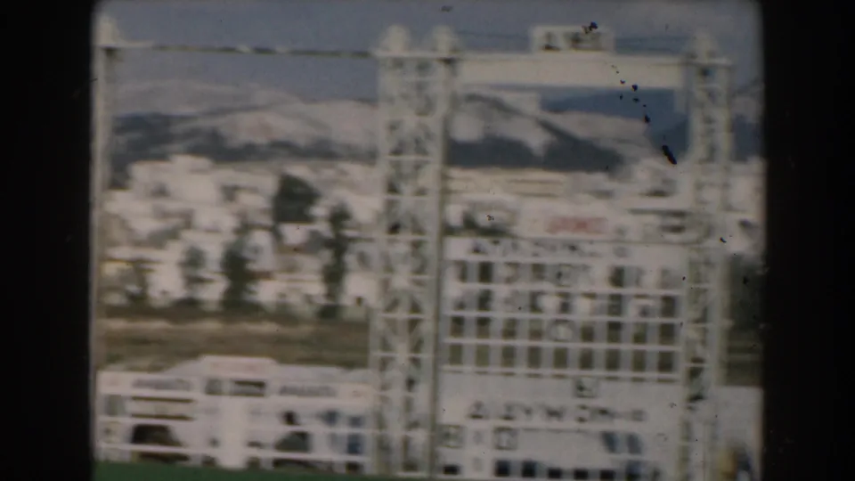 1962 Greece home-movie frame showing a passing scoreboard-like frame, a bus or coach, town buildings, and mountain slopes.