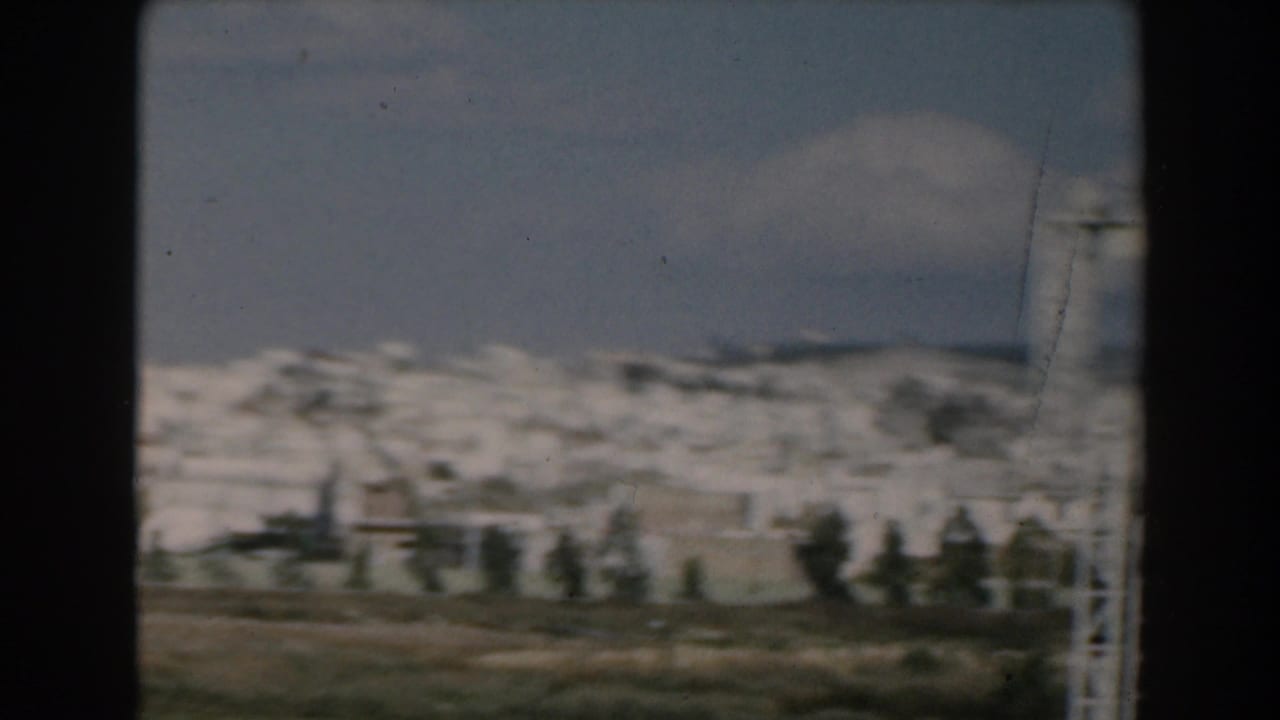 1962 Greece frame showing open land, distant white town buildings, trees, and a mountain backdrop, with a sign frame at the edge.