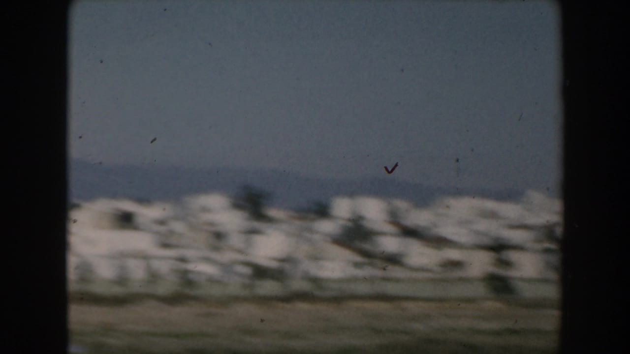 1962 Greece home-movie view of a small aircraft above white coastal buildings and distant mountains, seen from a moving vessel.