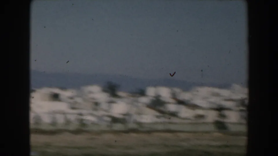 1962 Greece home-movie view of a small aircraft above white coastal buildings and distant mountains, seen from a moving vessel.