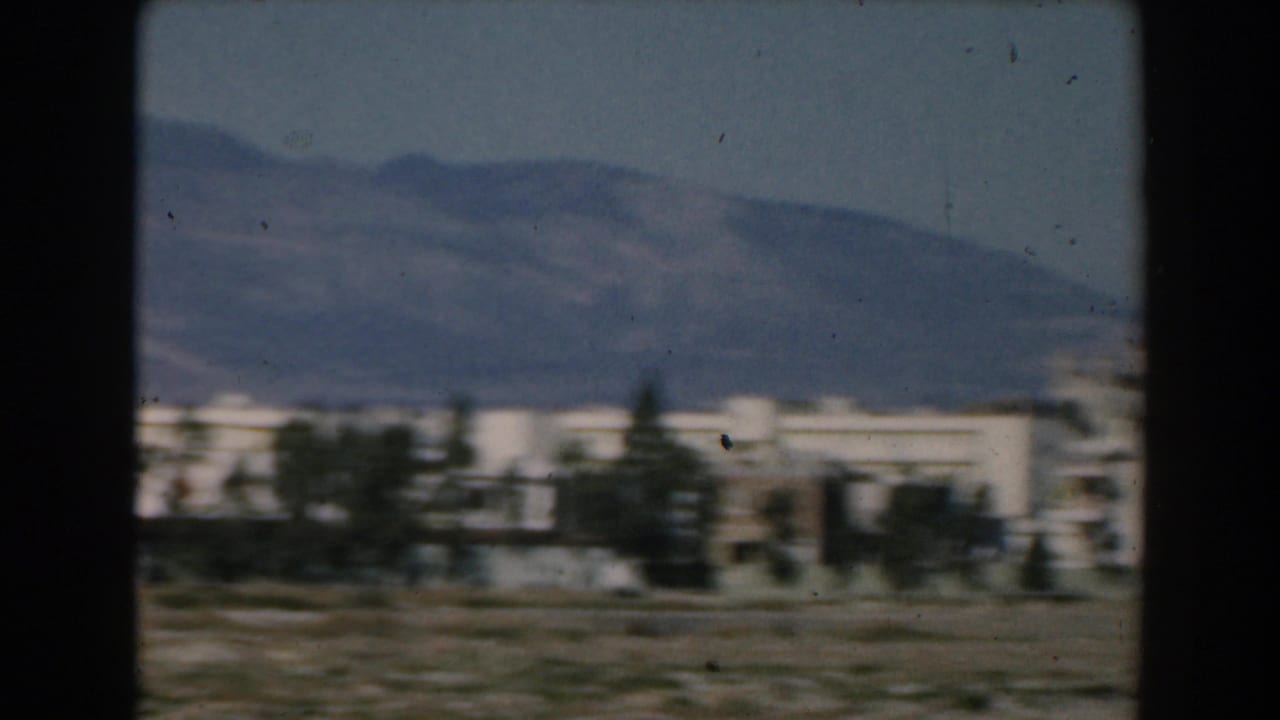Soft 8mm view of white low-rise buildings, scattered trees, and a mountain backdrop along the Greek coast in 1962.