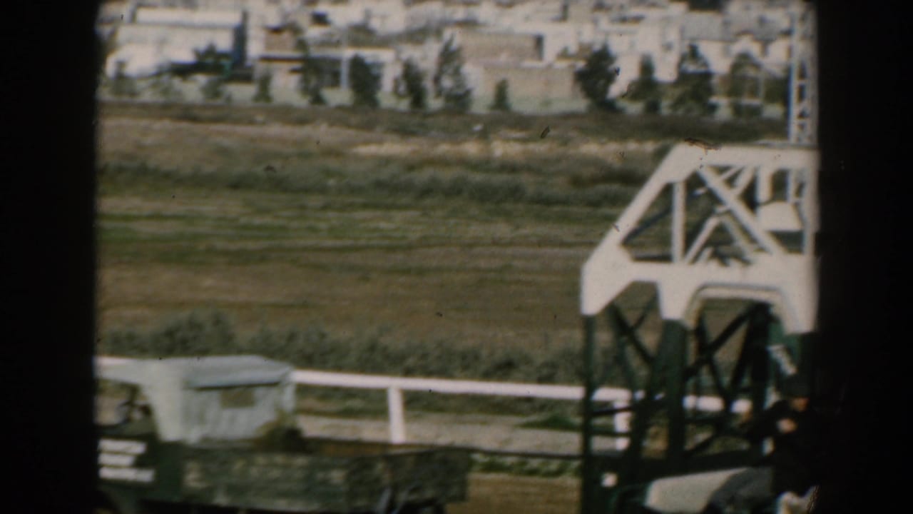 Moving shipboard view of open land, low buildings, trees, and a large white crane-like structure in 1962 Greece footage.