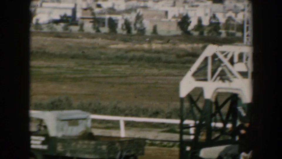 Moving shipboard view of open land, low buildings, trees, and a large white crane-like structure in 1962 Greece footage.