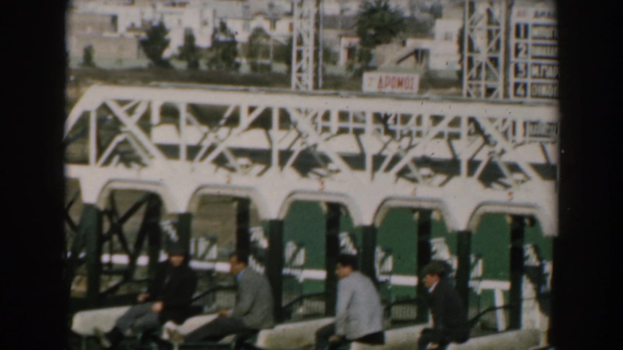 1962 Greece shipboard frame with passengers seated on deck as shoreline buildings and a white structure pass in the background.
