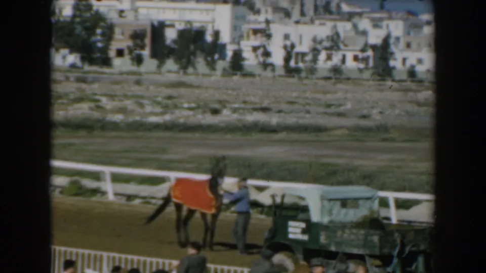 1962 Greece frame showing a horse led near the racetrack rail, with a green utility truck and city buildings in the background.