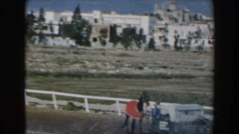 1962 Greece 8mm frame showing a horse and rider near the rail, with open ground, trees, and pale buildings in the background.