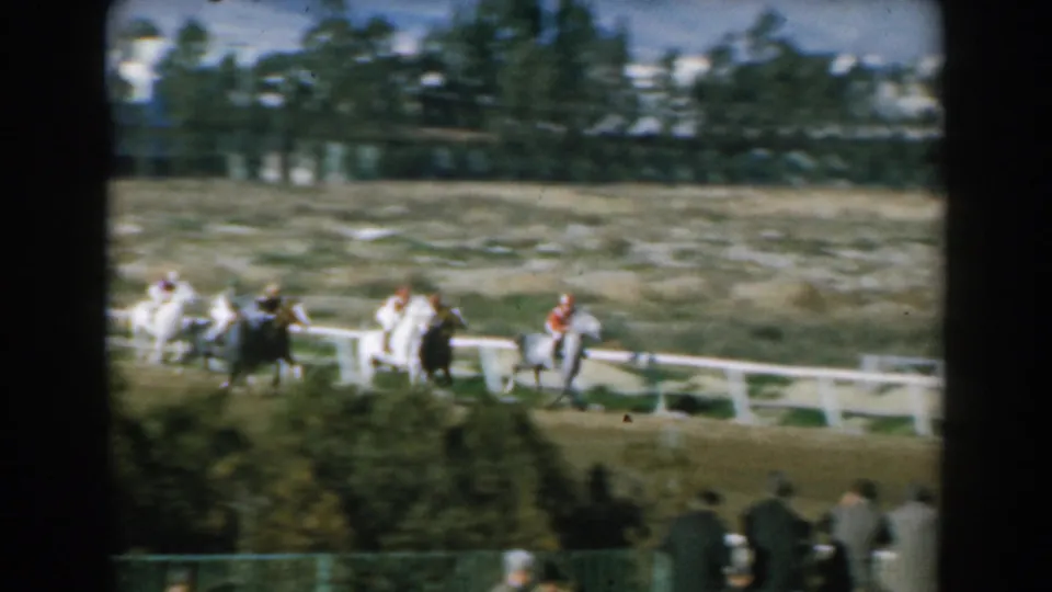 1962 Greece 8mm frame of racehorses and jockeys passing the rail, with spectators in the foreground and trees behind the course.