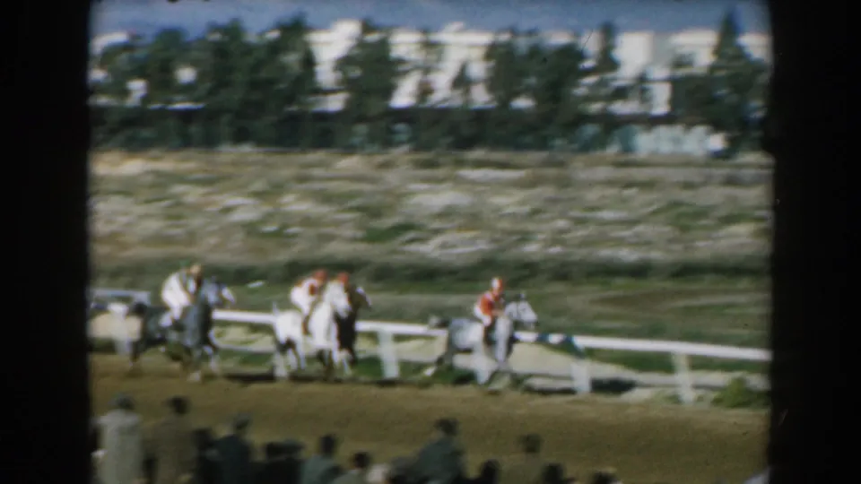 1962 Greece 8mm frame showing a pack of horses and jockeys along the track, with railings, trees, and town buildings in the distance.