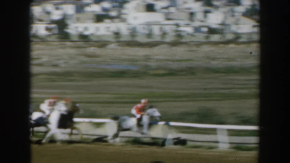 1962 Greece 8mm frame with motion-blurred horses and jockeys racing past white rails, with buildings and open ground beyond.