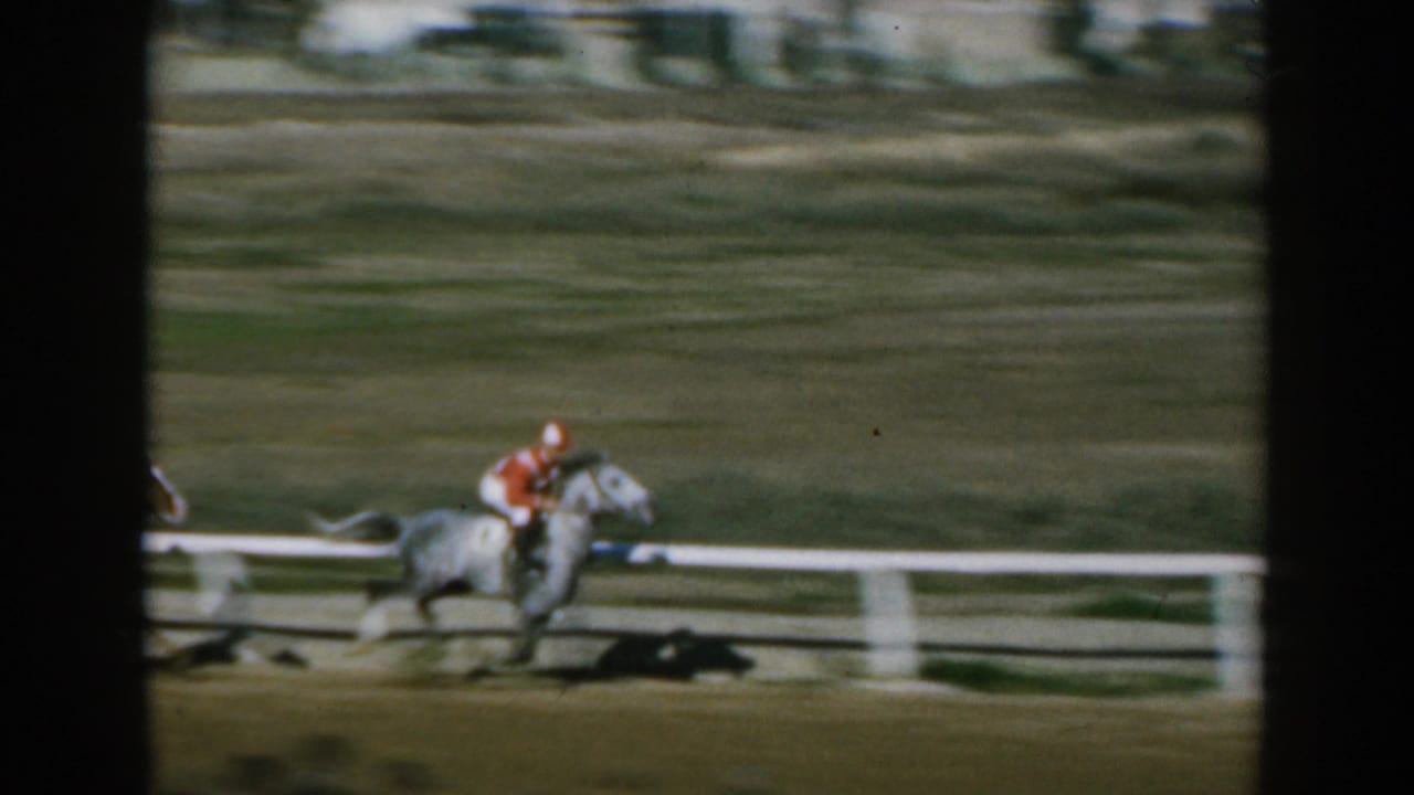 In 1962 Greece home-movie footage, a jockey in red rides a light-colored horse along a dirt racetrack, partly blurred by motion.