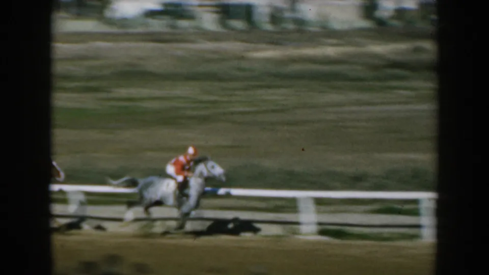 In 1962 Greece home-movie footage, a jockey in red rides a light-colored horse along a dirt racetrack, partly blurred by motion.