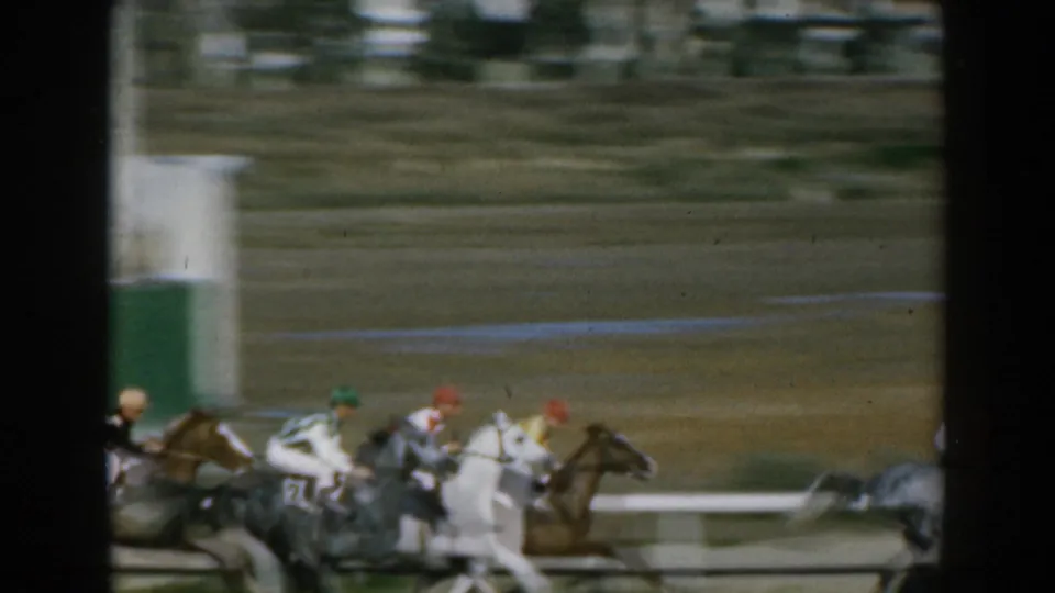 A group of jockeys in colored silks ride closely together on a Greek dirt racetrack in this 1962 8mm frame.