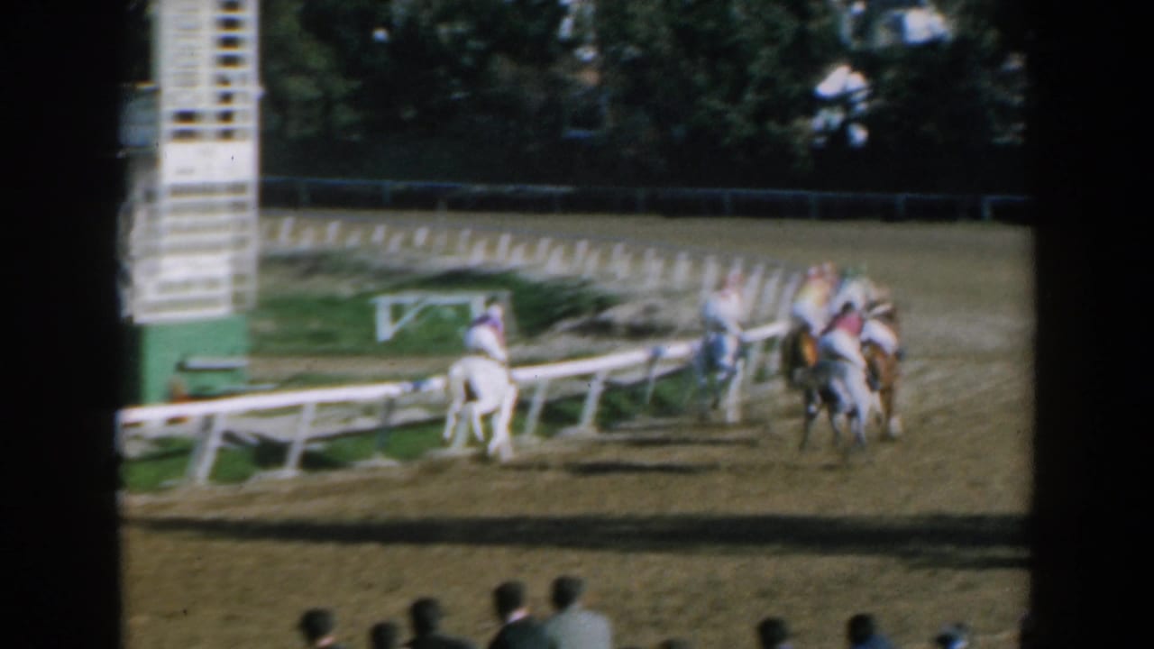 1962 Greece home-movie frame showing jockeys and horses rounding a dirt race track with spectators in the foreground.