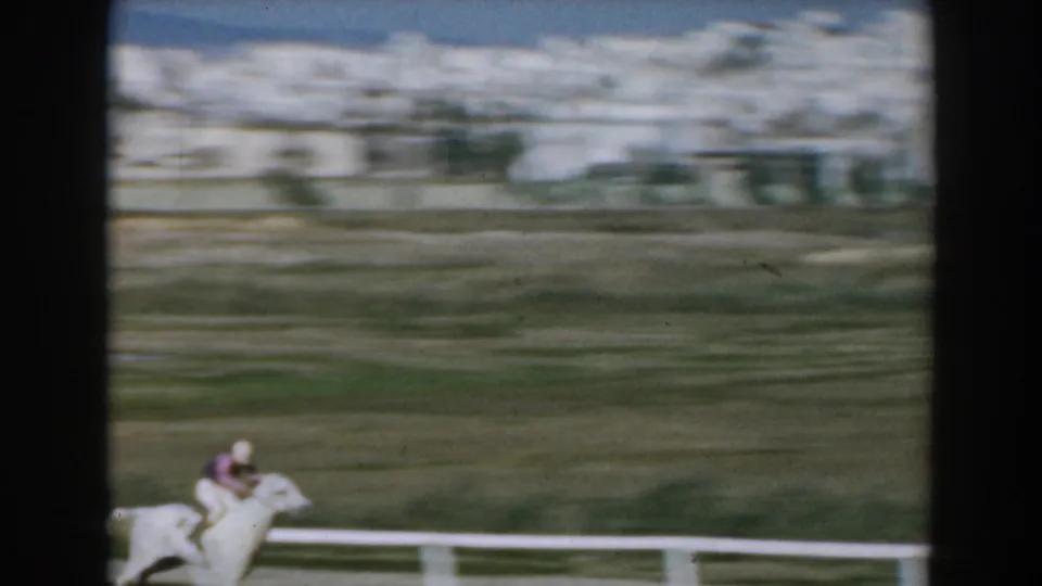 A jockey on a white horse gallops past the rail in a blurred 1962 Greece racecourse view.