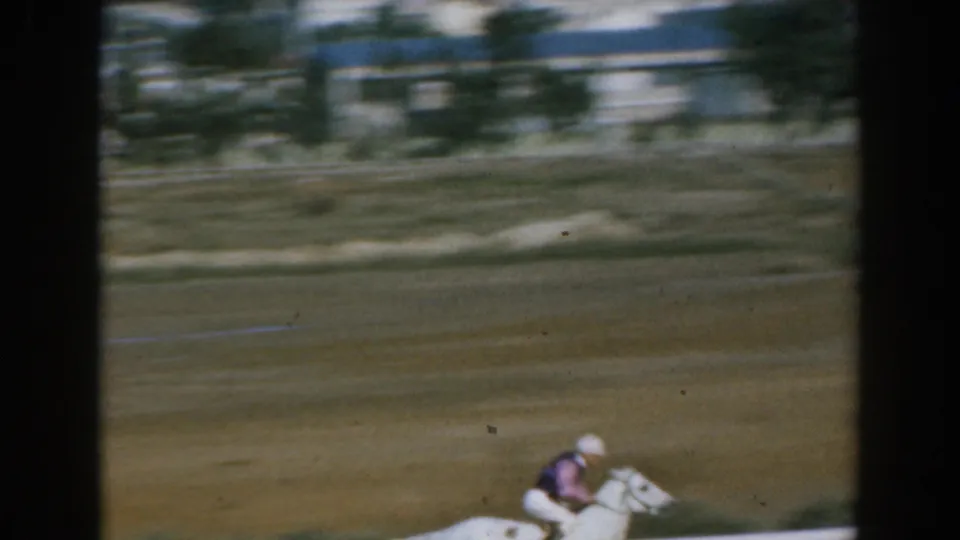 The white horse and rider continue along the dirt track, with blurred racecourse structures in the background.