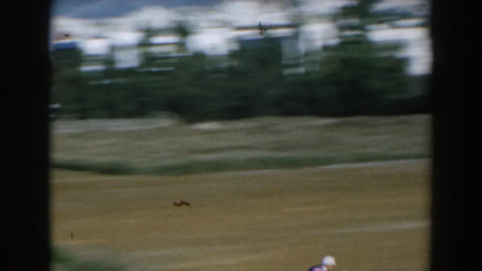 1962 Greece 8mm frame showing a blurred racetrack area through dark window bars, with trees and pale buildings in the distance.