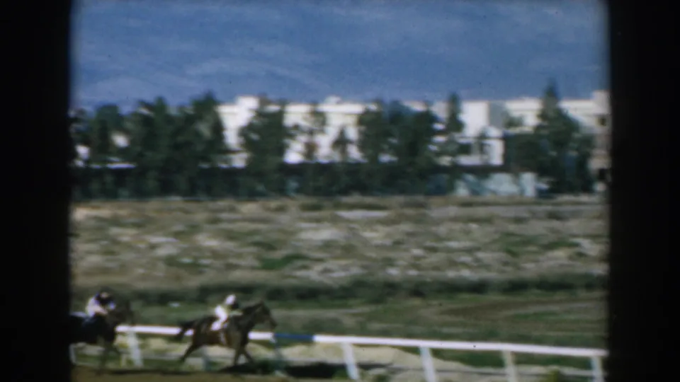 Two jockeys ride horses along a Greek racetrack in 1962, seen through dark framing with white buildings and trees behind them.