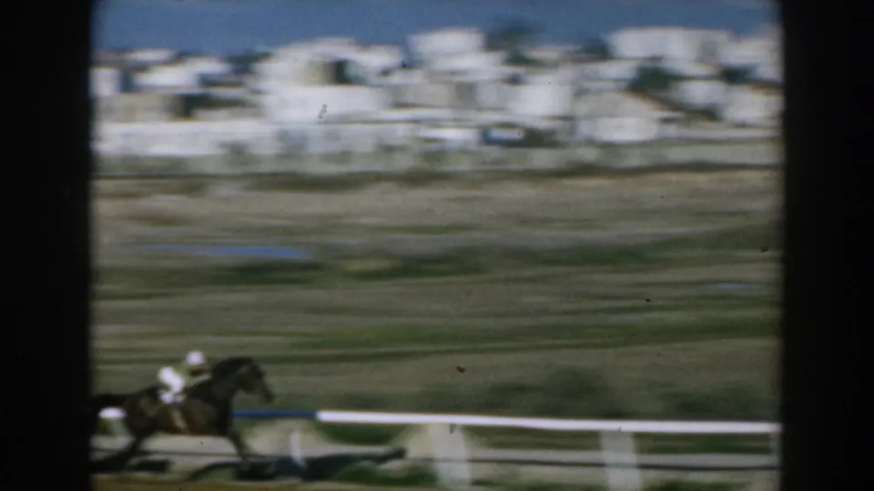 A jockey on horseback moves along the rail in 1962 Greece footage, with motion blur and distant white buildings behind the track.