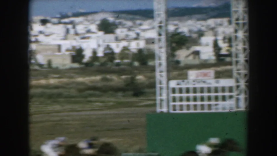 The closing frame shows the race continuing past a white track structure, with Greek buildings and trees beyond the course.