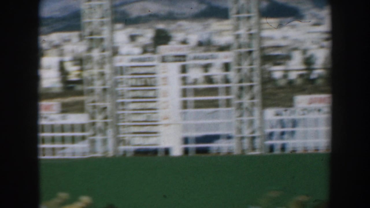 1962 Greece home-movie frame with a blurred white structure, green field, pale buildings, and mountains in the distance.