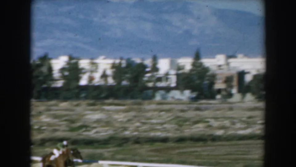 A horse and rider appear near a track rail in rural Greece, with white buildings, trees, and mountains in the background.