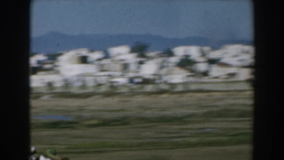 1962 Greek rural view with blurred white buildings, open land, distant mountains, and dark edges from the filming position.