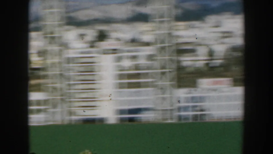 A blurred white track or scoreboard structure crosses the frame above a green field, with pale buildings and hills behind.