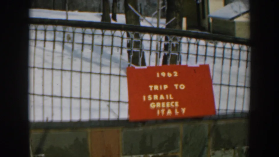 Red sign on a wire fence reads "1962 TRIP TO ISRAIL GREECE ITALY" in a snowy outdoor setting.