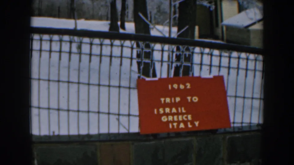 The 1962 travel sign remains centered on a fence, with snow, trees, and a small structure visible behind it.