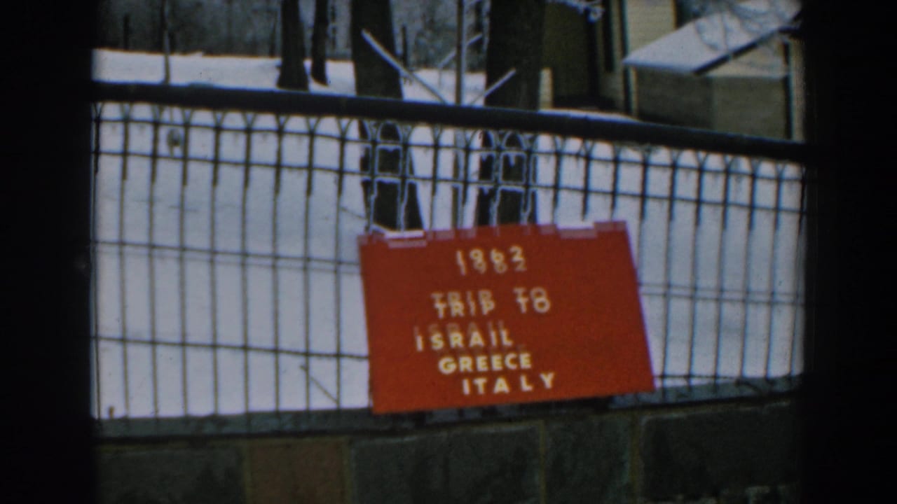 1962 home-movie frame showing a red travel board reading “TRIP TO ISRAIL GREECE ITALY” on a black fence.