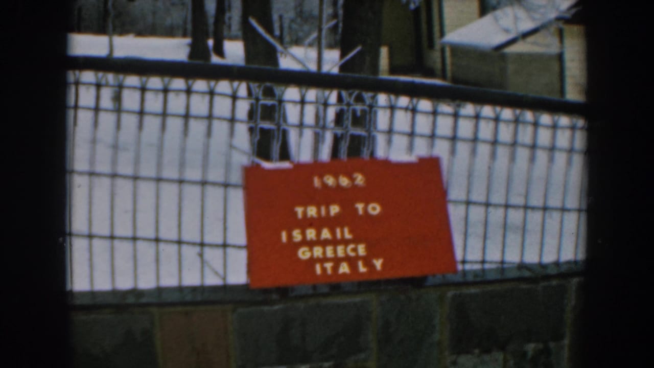 Static 1962 travel-sign frame with a red board listing Israil, Greece, and Italy behind a black fence.