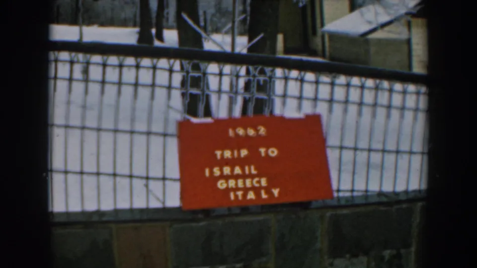 Static 1962 travel-sign frame with a red board listing Israil, Greece, and Italy behind a black fence.