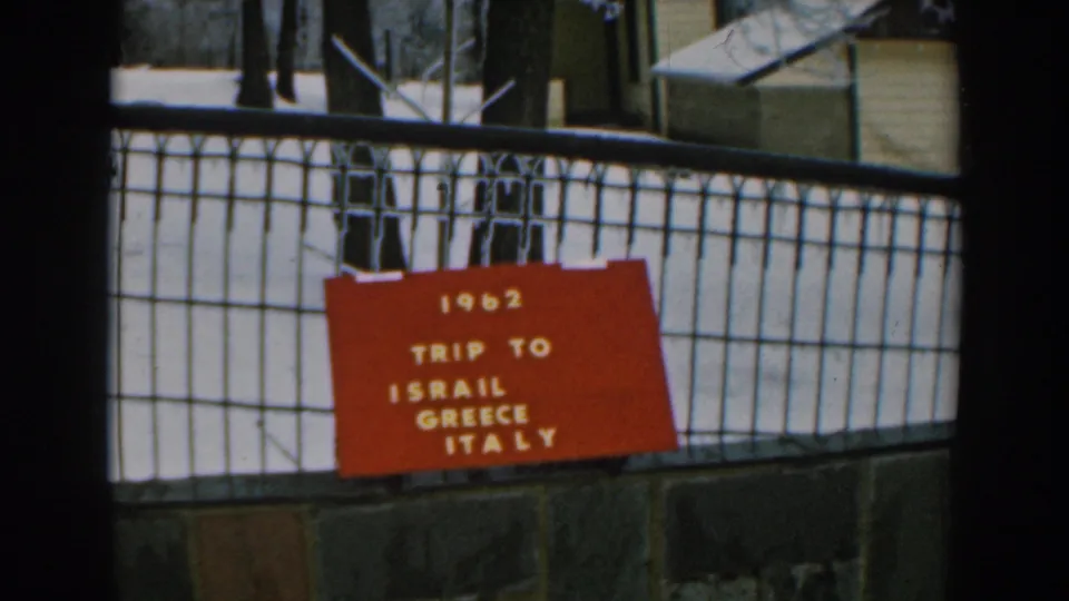 Final 1962 8mm frame holding on the red trip board, fence, snow, trees, and background structure.