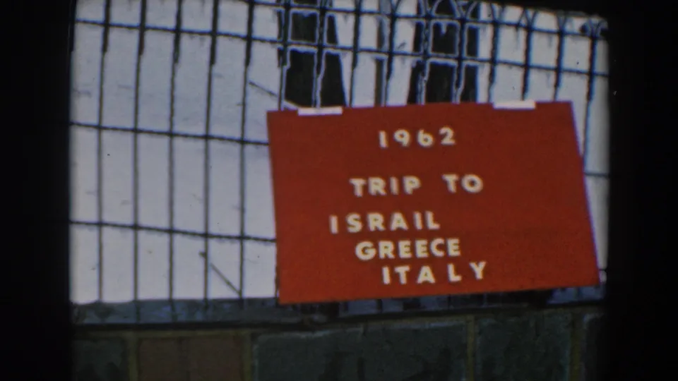 A red 1962 travel title card fills more of the frame, fixed to a metal grid with pale architecture behind it.