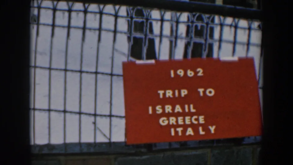 Centered 8mm frame of the red 1962 trip title card for Israel, Greece, and Italy, with gridwork and masonry behind it.
