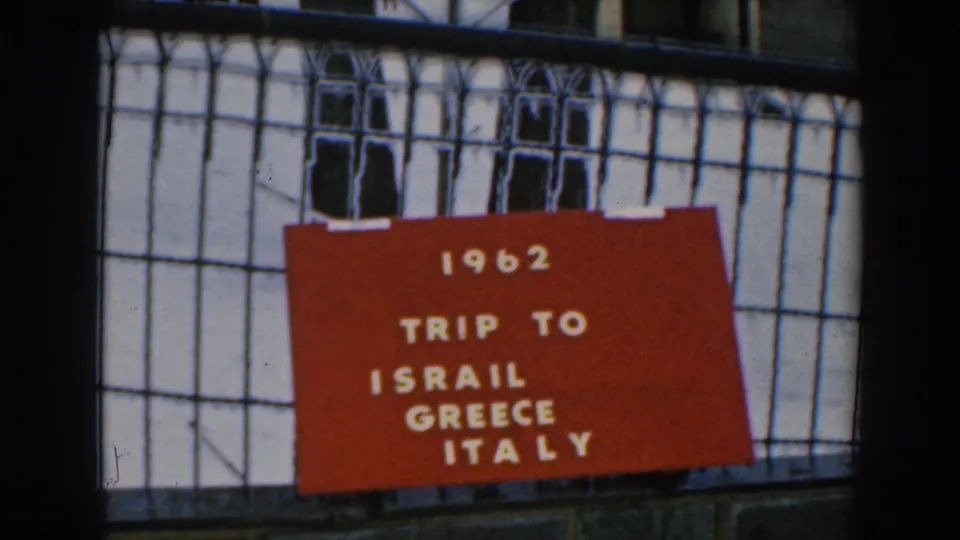 Final frame holds on the red 1962 travel card, with visible 8mm grain, soft focus, and the gridded exterior setting.