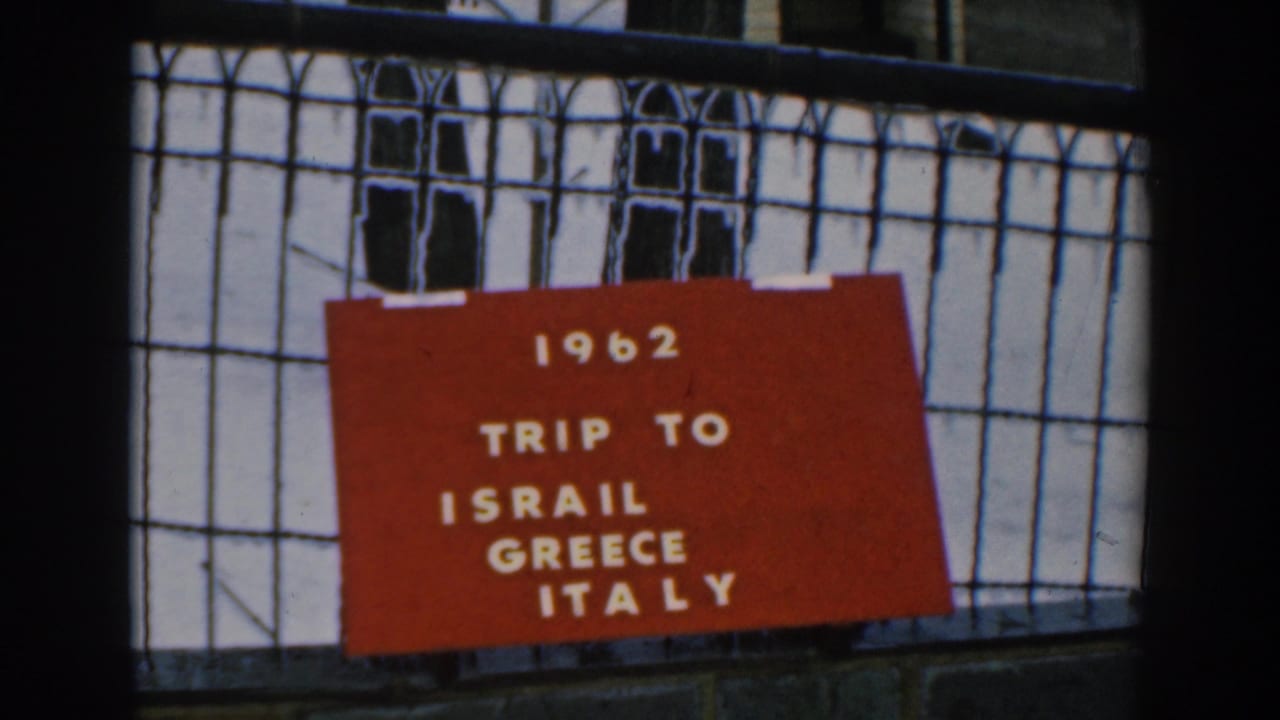 1962 home-movie frame showing a red card reading trip to Israel, Greece, Italy on a black fence outdoors.
