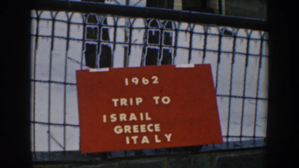 1962 home-movie frame showing a red card reading trip to Israel, Greece, Italy on a black fence outdoors.
