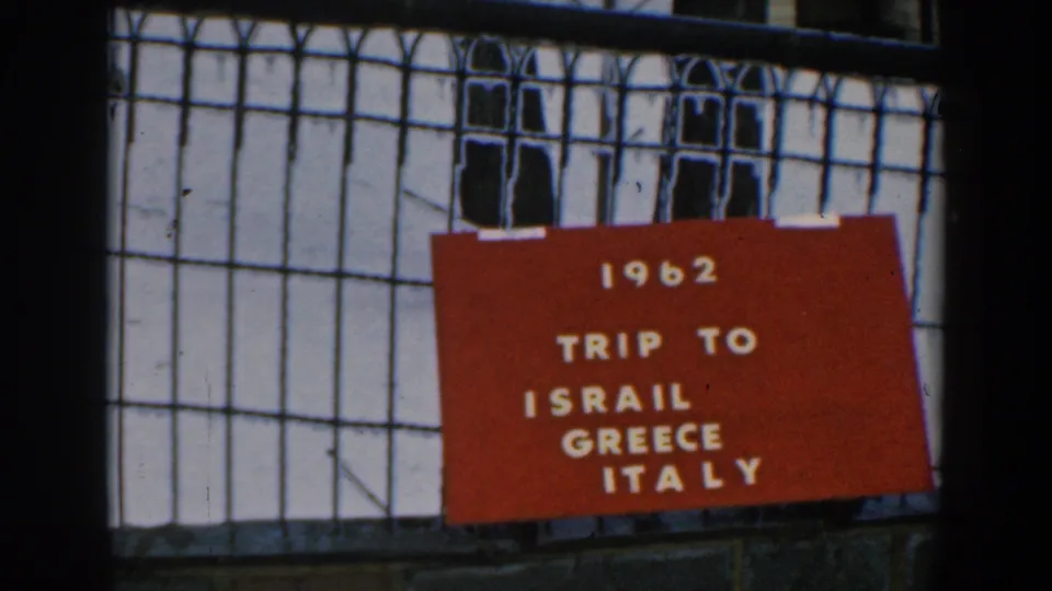 Close 8mm frame of a red 1962 travel title card on a fence, with the destination text visible.