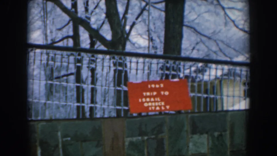 Wider 1962 home-movie view of the red trip card on a fence, with bare trees and a stone wall behind.