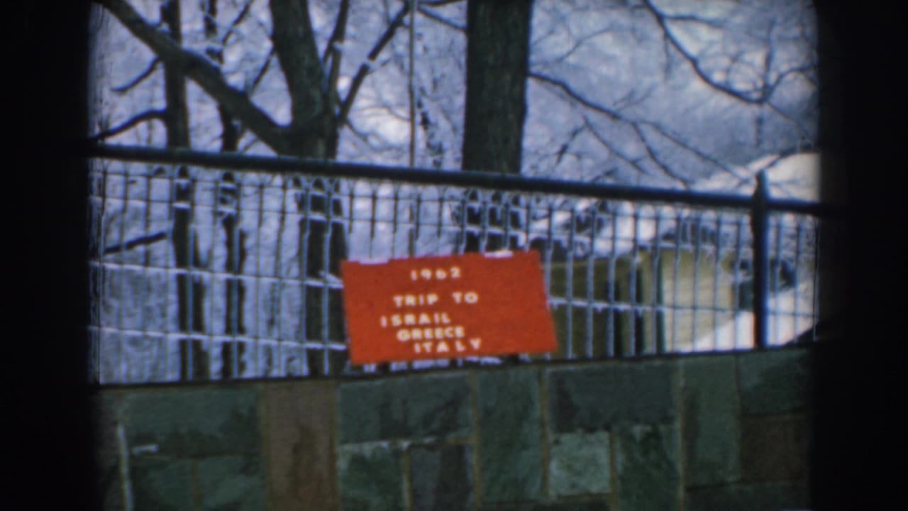 1962 Greece travel-film frame showing a red “TRIP TO ISRAEL GREECE ITALY” sign behind a wire fence and stone wall.